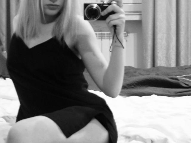 Schwarz-weißes Foto von Tserera mit blonden Haaren, die ein eng anliegendes schwarzes Kleid trägt und auf einem Bett sitzt. Sie hält eine Kamera, wobei ihr nacktes Oberschenkel und ein Teil ihres Gesäßes zu sehen sind..
