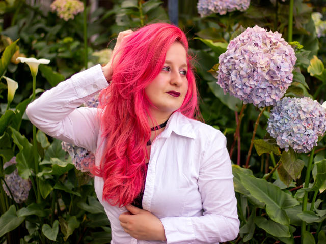 VioletaMora with bright pink hair, white shirt, and dark lipstick, hand in hair, looking to the side, surrounded by purple hydrangeas, green leaves, and white flowers.