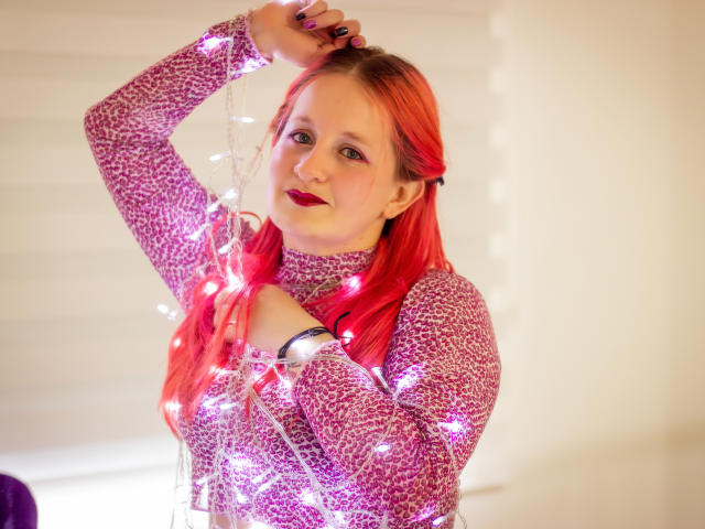 VioletaMora with red hair, wearing a purple sparkly sweater, holding glowing Christmas lights across her chest and arm, smiling with red lipstick.