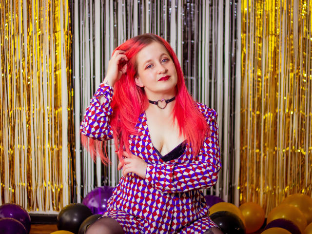 VioletaMora with bright red hair, wearing a checkered dress with a deep V, black choker, and black bra, sitting on balloons, hand in hair, smiling. Boobs visible.