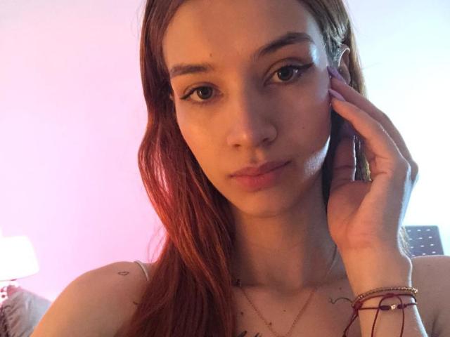 Greta Harris tem cabelos ruivos longos, olhos castanhos e um sorriso discreto. Ela toca o rosto com uma mão, mostrando uma tatuagem no ombro e uma pulseira no pulso..