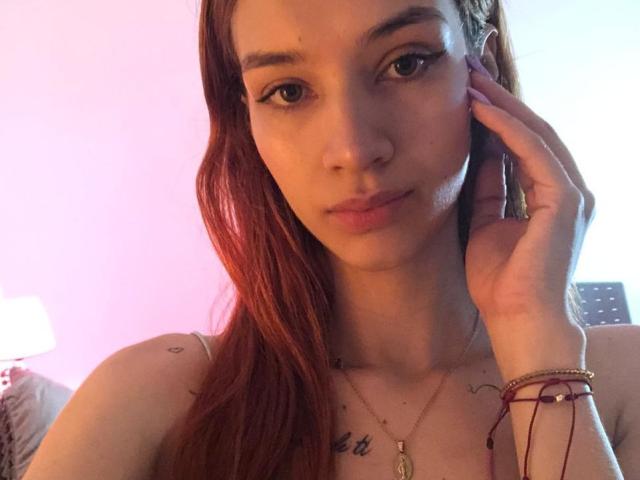 Greta Harris, com cabelo ruivo, segurando a bochecha, tem seios pequenos, tatuagens no peito, olhos castanhos, está usando um colar e pulseiras, em um quarto com iluminação suave..