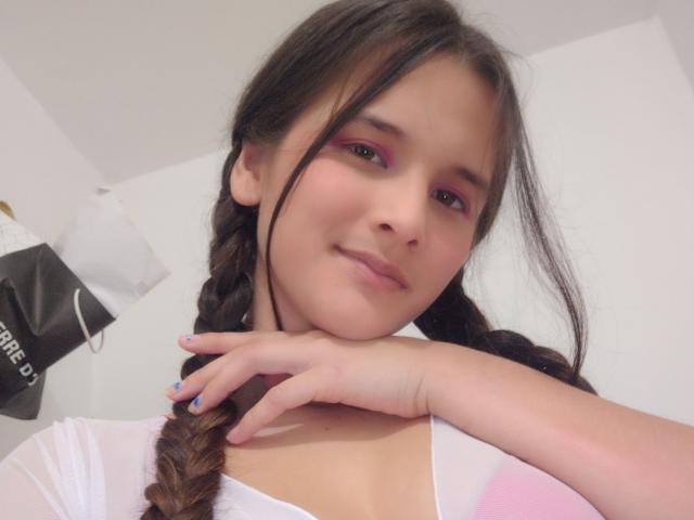 AllysonAndMax met gevlochten bruin haar, roze oogschaduw, een witte top, hand op de nek, blauwe nagels, een lichte glimlach, bleke huid, zacht licht, een schattig gezicht, kleine borsten, een casual selfie..