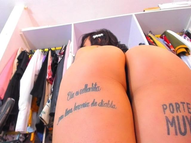 YeiSela's ass is tattooed with text on both thighs, her tan skin is visible, and she's standing in front of a clothes rack with her back to the camera.