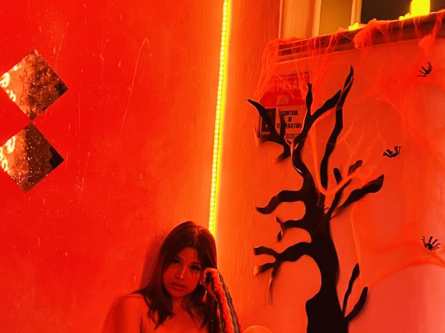 Red-lit photo of ShantalSummer with long brown hair, medium tits, and a slight smile, holding a phone against a wall with black tree and neon strip.