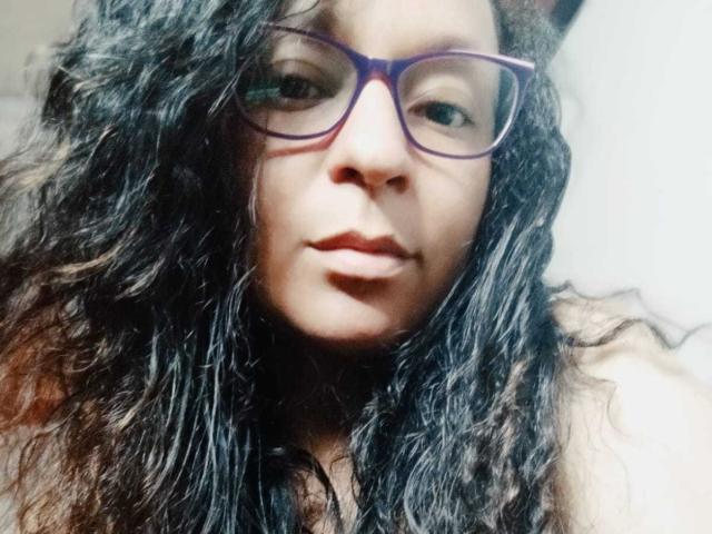 Angelonix with curly black hair, wearing purple glasses, has a serious expression and is looking at the camera, showing her nose and slightly parted lips.