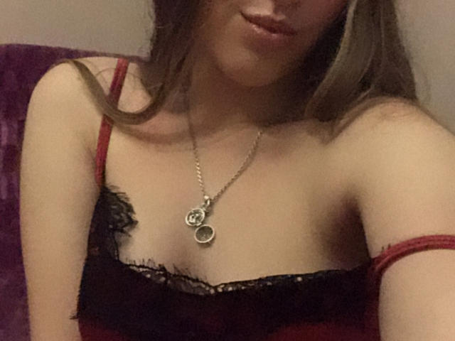 Mulher com cabelo castanho, vestindo lingerie de renda vermelha e preta, colar com dois pingentes circulares, sorriso discreto, decote visível, alça vermelha parcialmente fora do ombro..