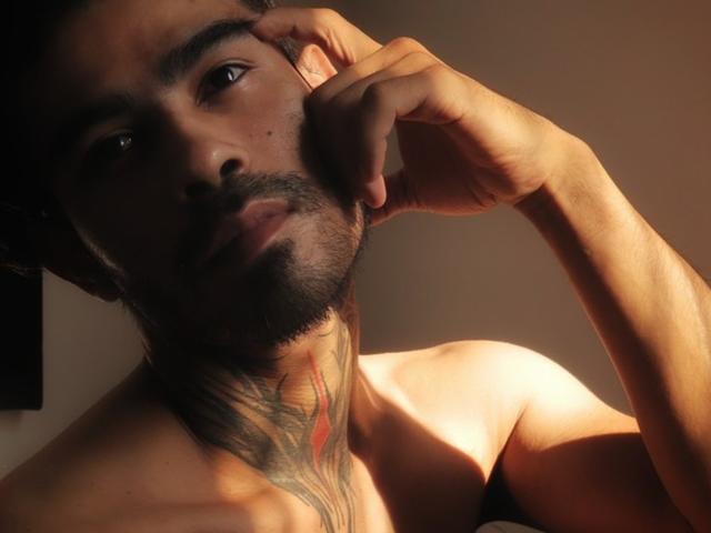 Stefanoeverett hat dunkles Haar, einen Bart und ein Tattoo im Nacken, das eine Hand zeigt, die sein Ohr berührt. Er ist ohne Hemd und wird von warmem Sonnenlicht beleuchtet. Sein Gesichtsausdruck ist leicht nachdenklich..