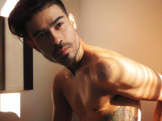 Stefanoeverett senza camicia, con capelli scuri, barba e tatuaggi, piegato in avanti, con la luce del sole che proietta ombre sul suo petto, occhi marroni, leggero sorriso, che tiene il braccio sinistro..