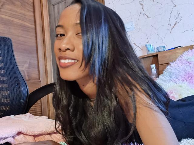 LunaCuty, con cabello negro largo, sonriendo y mostrando dientes blancos, vestida con una blusa negra, sentada en una habitación de madera, con una manta rosa en primer plano y una silla negra a la izquierda..