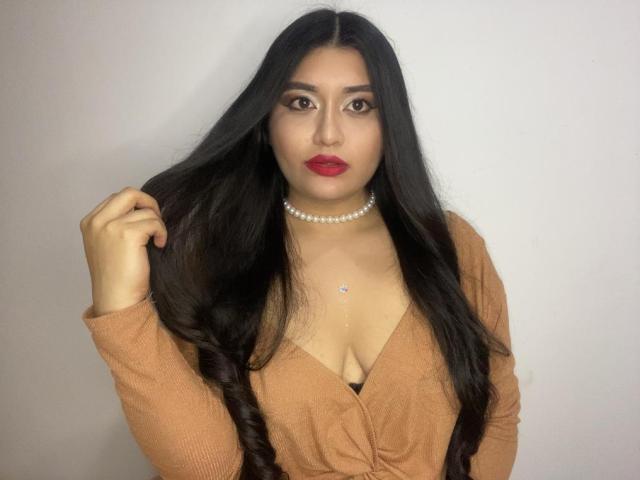 MiaDuboisX with long black hair, red lipstick, pearl necklace, and tan cardigan, holding hair, showing cleavage, wearing a black bra, looking at camera.