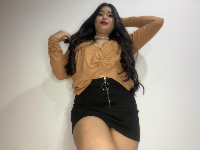 MiaDuboisX with long black hair, red lipstick, brown top, black skirt with zipper, and pearl necklace, standing with arms raised, showing her tits and ass.