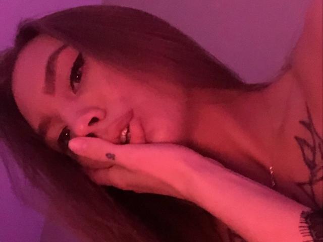 LexiLee's face in a pinkish-purple light, biting her finger with a small black tattoo on her hand, showing a tattoo on her chest. Her long brown hair is down.