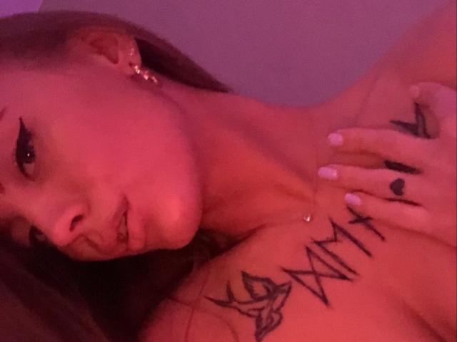 LexiLee with pinkish-red lighting, showing tattooed shoulder, black XIII and rose tattoos, black ringed finger, white nail, ear piercing, and parted lips, lying down.