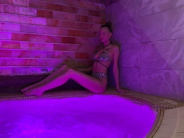 LexiLee with small tits and a leopard print bikini, sitting by a purple-lit hot tub, legs crossed, arms resting, brown hair in a bun, looking relaxed.