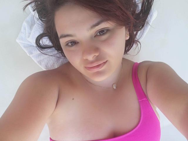 KatherinLatin deitada, olhando para a câmera com um leve sorriso, vestindo uma regata rosa, mostrando seus seios, com cabelo castanho e uma toalha branca sob a cabeça..