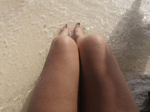 Les cuisses hâlées de katherinLatin, parsemées de gouttes d'eau, genoux serrés, pieds dans une eau peu profonde et claire, orteils légèrement écartés, fond sableux visible, la lumière du soleil se reflétant sur la peau..