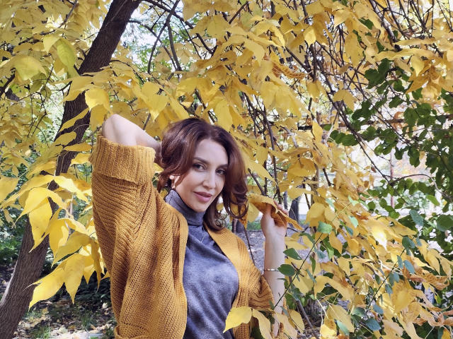 SimoniGrace com cabelo castanho ondulado, casaco amarelo, gola alta cinza, segurando uma folha amarela, sorrindo, floresta de outono, mão no cabelo, seios pequenos, fofa, luz natural, sorriso suave..