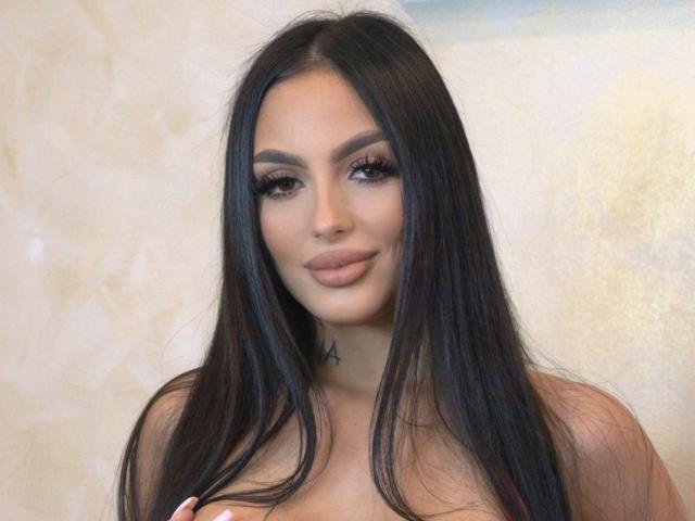 KailynX ha lunghi capelli neri lisci, sopracciglia folte, ciglia spesse e sta sorridendo con le labbra piene. Ha un piccolo tatuaggio vicino al collo ed è senza top..