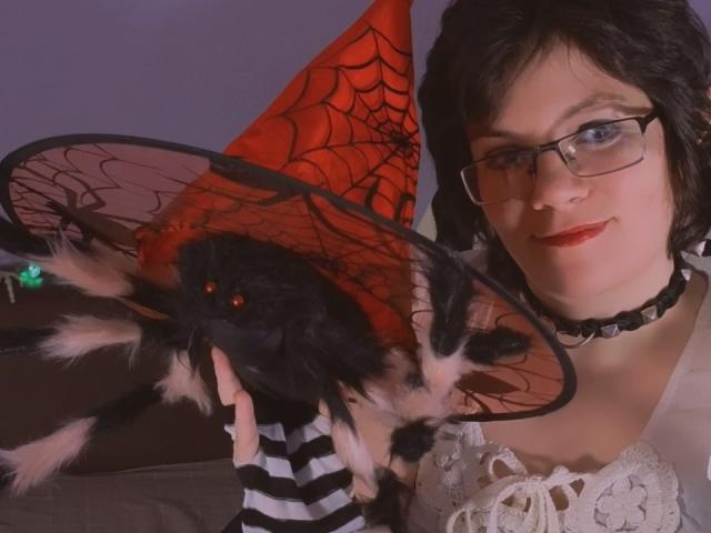 EverlyRays with black hair, glasses, and red lipstick, holding a black furry spider with red eyes, striped socks, and a red spiderweb hat. Choker around her neck.