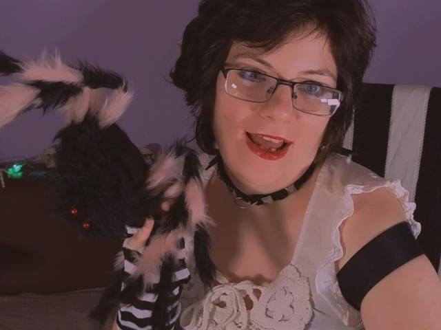 EverlyRays with short black hair, glasses, and red lipstick, wearing a white lace top, black choker, and arm band, holding a fluffy black cat with pink ears.