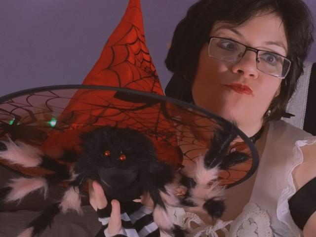 EverlyRays with glasses, black hair, and red lipstick, holding a black and pink fuzzy spider with red eyes and a web-patterned red hat.