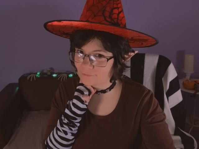 EverlyRays, wearing a red witch hat with black webbing, striped sleeves, and a choker, has short black hair, glasses, and a brown shirt, with a thoughtful expression.
