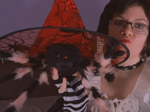 EverlyRays with glasses, pink lipstick, black choker, white lace top, holding a black spider with pink and black fuzzy legs, and red eyes, in a dimly lit room.