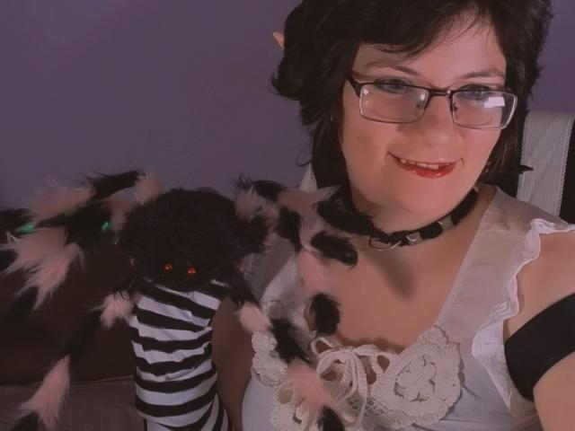 EverlyRays with black hair, glasses, and red lipstick, wearing a white lace top and black-and-white striped arm, holding a fluffy black spider toy with pink accents.