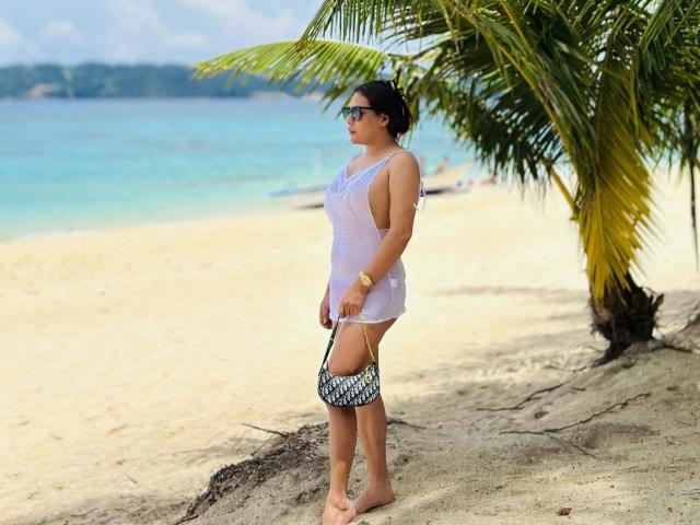 TikolyAma, con un seno di media grandezza, indossa un abito bianco trasparente, una borsa a motivi bianchi e neri, è in piedi su una spiaggia sabbiosa, con una palma a destra, indossa occhiali da sole e guarda di lato..