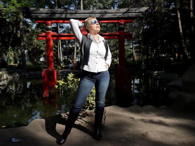 Catherine, een volwassen vrouw met kort blond haar, blauwe ogen, een wit shirt, een zwarte vest, blauwe jeans en zwarte laarzen, staat voor een rode torii-poort, met haar hand op haar hoofd, in het zonlicht, met een bos op de achtergrond..