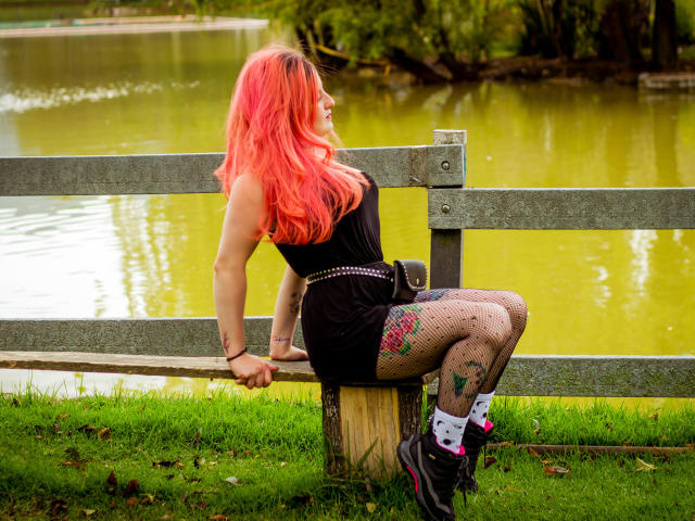 VioletaMora with pink hair, black dress, fishnet stockings, tattoos, and white socks with pink hearts, sitting on wooden post by green-tinged pond, leaning on gray fence.
