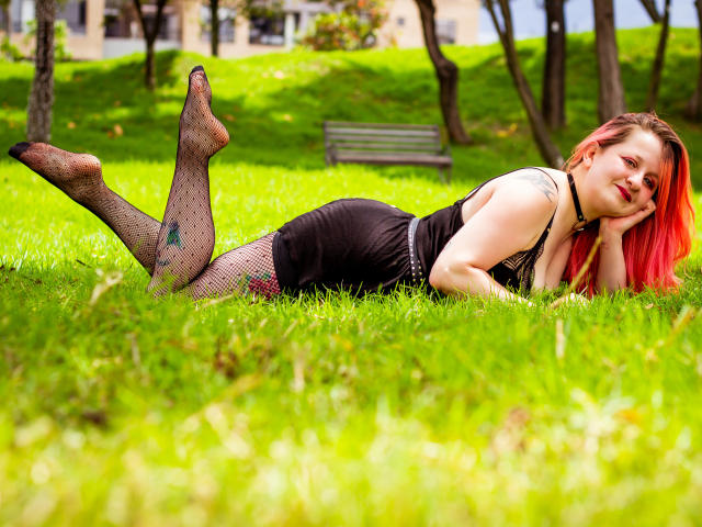 VioletaMora lies on grass, wearing a black dress and fishnet stockings, her red hair and fair skin contrasting with the green. Her ass is up, feet in the air.