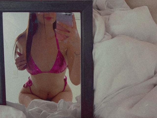 Nikole Foster usando um biquíni rosa, mostrando seus seios e nádegas, tirando uma selfie no espelho com o celular, com seu longo cabelo castanho parcialmente visível, em uma cama com lençóis brancos..