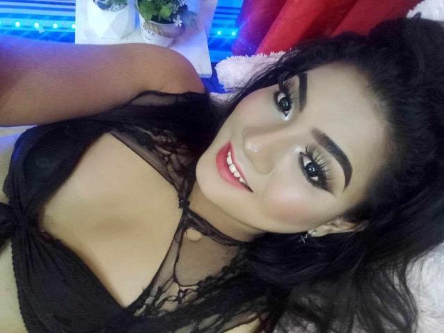 QueenMilker, com cabelo escuro, olhos castanhos e lábios cheios, vestindo um sutiã de renda preto, mostrando seus seios, deitada em uma cama, sorrindo para a câmera..