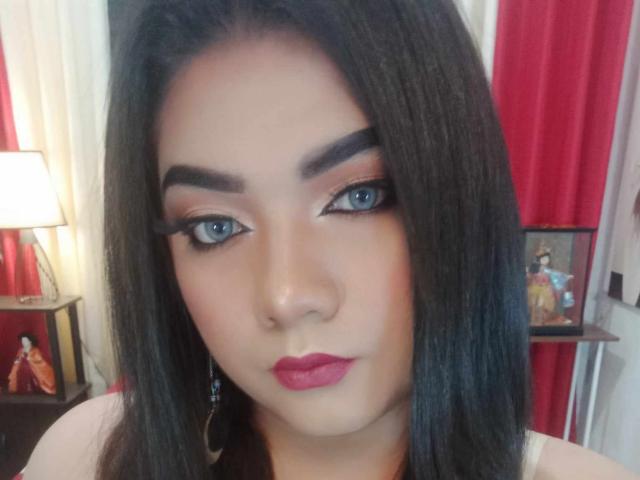 QueenMilker hat langes, schwarzes Haar, blaue Augen, dicke Augenbrauen und roten Lippenstift. Sie trägt ein weißes Oberteil und hat einen Nasenring. Ihr Make-up ist auffällig und sexy..