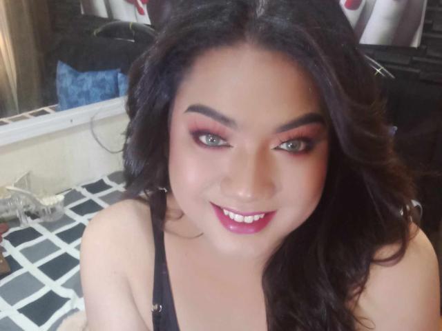 QueenMilker, com cabelos escuros e ondulados, sombra vermelha e batom rosa, sorrindo para a câmera, mostrando seus dentes brancos, vestindo uma blusa preta e parecendo adorável..