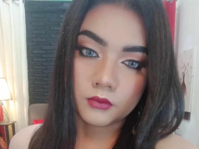 QueenMilker mit dunkelbraunen Haaren, blauen Augen und rosafarbenem Lippenstift hat einen ernsten Gesichtsausdruck und trägt Make-up mit auffälligen Augenbrauen und langen Wimpern, in einem sanft beleuchteten Raum..