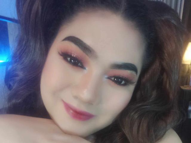QueenMilker hat welliges, braunes Haar, dunkle Augenbrauen und rosigen Lidschatten mit langen Wimpern. Sie lächelt mit rosafarbenem Lippenstift und legt ihr Kinn in ihre Hand..