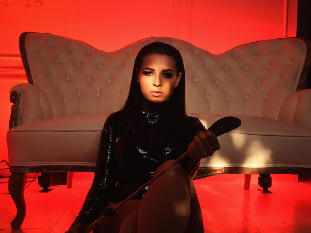 KayaOcean with black latex outfit, collar, and long hair, holding a black whip, sitting in front of a tufted beige sofa, illuminated by red lighting.