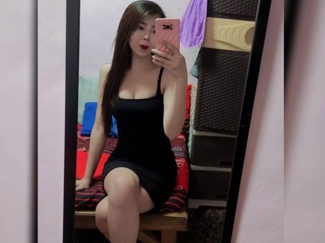 MonaHugeSelfSucker in un abito nero, aderente, con spalline sottili, seduta, che si scatta un selfie allo specchio, mostrando i suoi seni e il sedere, tenendo un telefono rosa, con lunghi capelli castani..