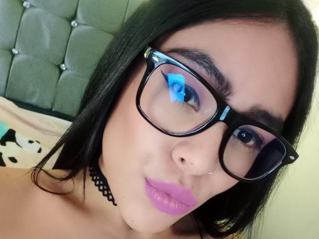 StellaBeckker with black glasses, purple eyeliner, blue reflection, pink lipstick, black choker, and long black hair, showing her cute face and small nose. Boobs not visible.