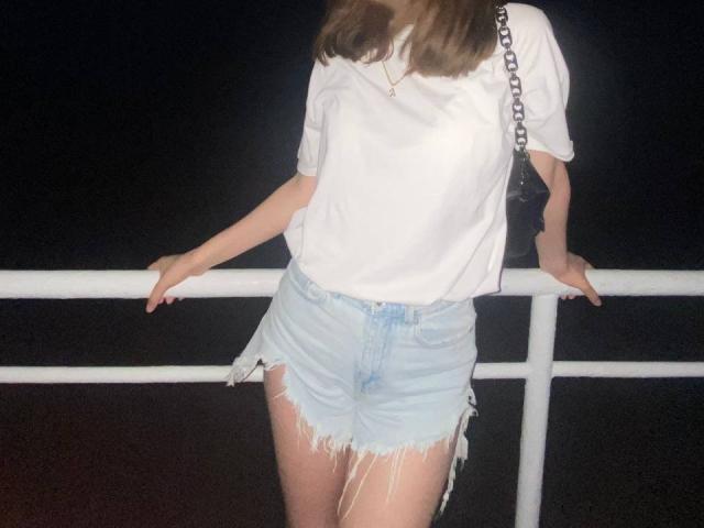 Tserera with frayed denim shorts, white tee, chain bag, leaning on white rail, hands on rail, head out of frame, dark background, visible thighs, long brown hair.
