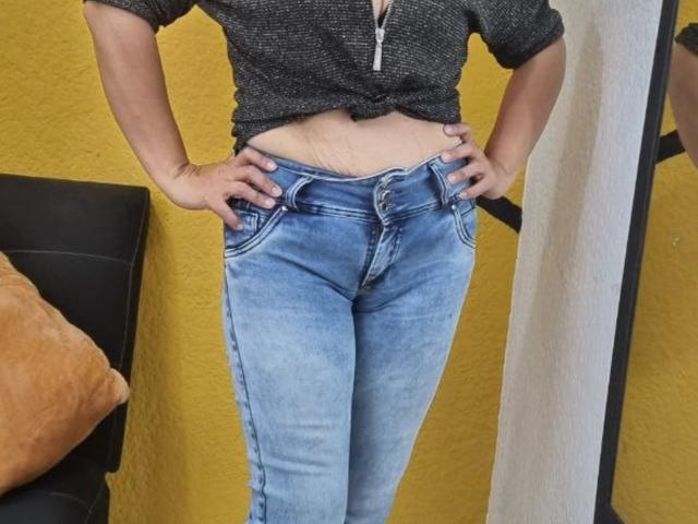 CatalinaJH hatte die Hände an der Hüfte, trug blaue Jeans mit einer sichtbaren Bräunungslinie und eine schwarze, hochgebundene, gefleckte Bluse, die ihren leicht gewölbten, nackten Unterbauch zeigte..