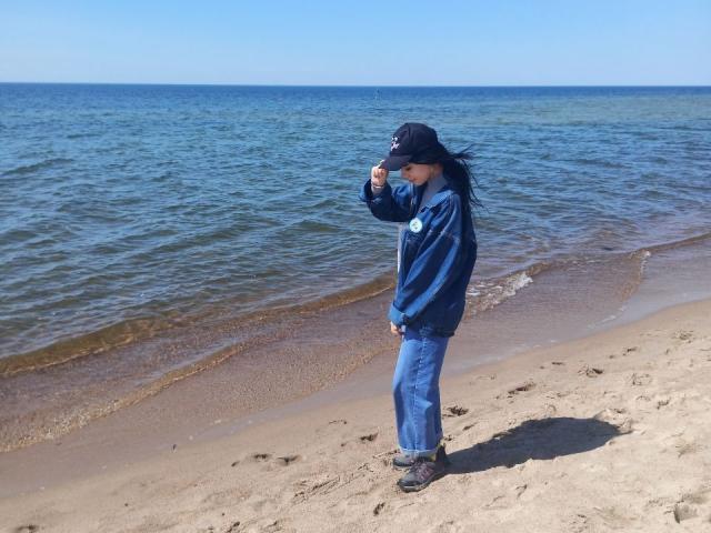 Zanoza, die eine blaue Jacke und Mütze trägt, steht auf einem sandigen Strand, hält ihre Mütze mit ihrer rechten Hand und hat langes, schwarzes Haar, das im Wind weht..