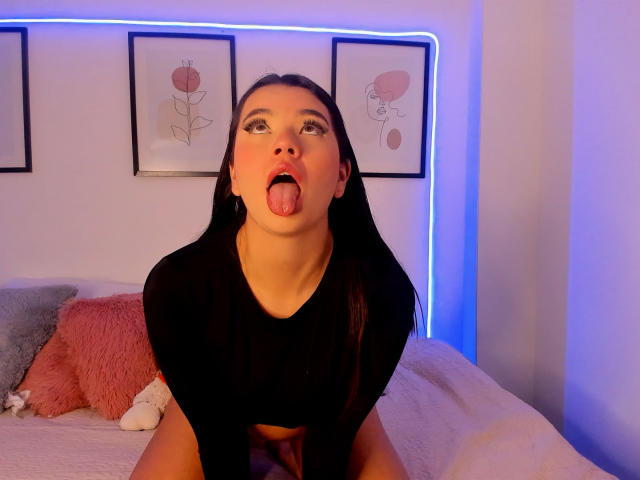 KatherineSummer, black long hair, tongue out, black crop top, bed, blue neon light, framed art, pink and gray pillows, looking up, cute, sexy, slightly messy.