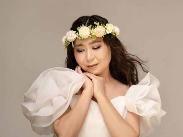 JeniBlue com uma coroa de flores, vestido branco com mangas bufantes, sorriso suave, mãos perto do rosto, cabelo castanho ondulado, pele clara, rosas rosa e brancas, expressão gentil, fundo neutro..