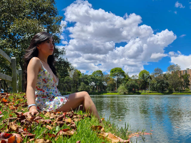 JainaRogers with dark hair, floral dress, sitting by a lake, legs spread, looking right, blue sky, fluffy clouds, autumn leaves, grass, white fence, trees, wrist bracelet.