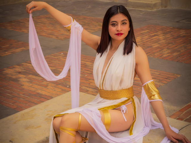 Samantha Smith, mit schulterlangem, schwarzem Haar, trägt ein weißes und goldenes Kleid im griechischen Stil, hält ein rosafarbenes Band, zeigt ihre nackten Oberschenkel und goldene Armreifen und sitzt auf einem Ziegelboden..
