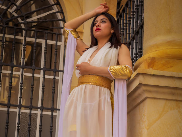 Samantha Smith, con cabello oscuro, lápiz labial rojo y brazaletes dorados, vestida con un vestido blanco transparente con una banda dorada, de pie junto a una ventana de hierro negro, con el brazo levantado..